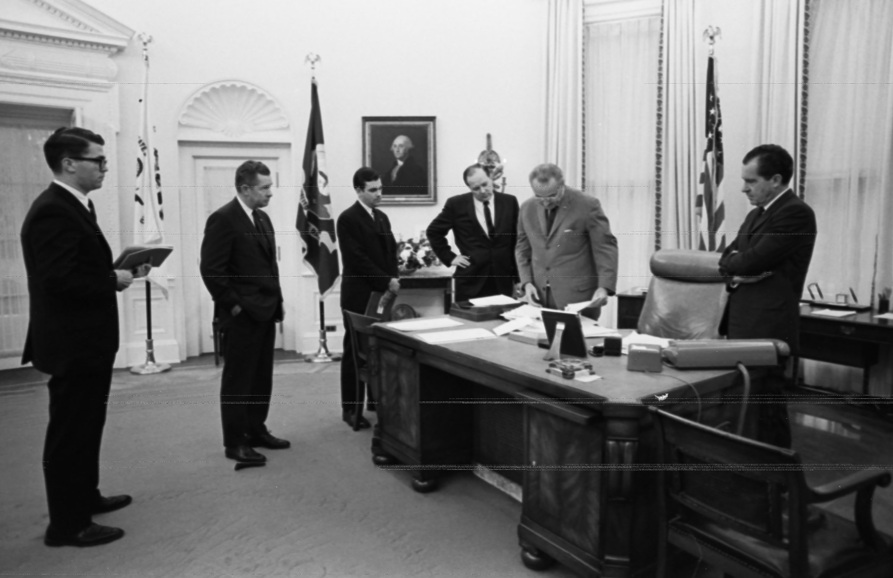 LBJ, center right, looks at papers at the desk of the Oval Office, while several men, including Richard Nixon, look on. 